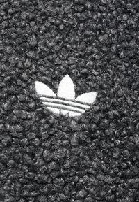 Γκρίζο υφασμένο ύφασμα με ένα λευκό κεντημένο λογότυπο τριών φύλλων. Η επιφάνεια έχει μια καμπυλωτή, κυκλική σχεδίαση, προσθέτοντας οπτικό ενδιαφέρον.