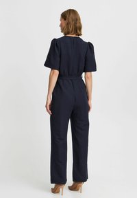 Tuta navy con maniche a sbuffo corte, tessuto strutturato, vita fitted e pantaloni larghi. Indossata con tacchi, mostra un dettaglio con zip sul retro.
