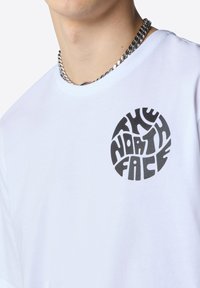 Biała bawełniana koszulka z krótkimi rękawami i okrągłym, czarnym graficznym logo z napisem "THE NORTH FACE" z przodu. Gładka faktura.