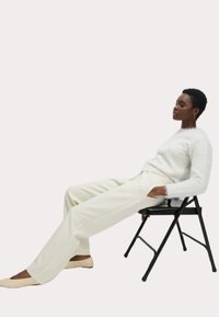 Un pull moelleux blanc, un pantalon large crème et des ballerines beiges. Assise sur une chaise pliante noire avec une main reposant sur la jambe.