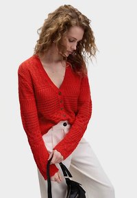 Červený pletený kardigan s výstřihem do V, zapín�áním na knoflíky a texturovanými vzory. Nošený s světlými kalhotami a černou kabelkou.