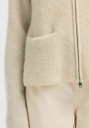 Gros plan sur un pull à fermeture éclair en tricot beige avec une poche frontale, porté avec un pantalon de couleur claire.