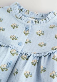 Ljusblå klänning med blommönster, volangkrage och knappar. Tillverkad av textilier med strukturerad yta och broderade blommor i rosa och blått.