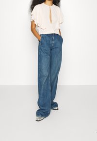 Jasnoróżowa bluzka z falbaniastymi rękawami i dekoltem w kształcie dziurki, w połączeniu z niebieskimi dżinsami o szerokich nogawkach oraz srebrnymi butami na obcasie.