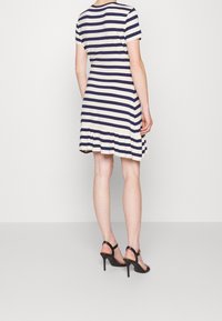 Pruhované krátké šaty s krátkým rukávem v námořnické modři a smetanově bílé, s rozšířeným lemem a jemnou látkou. Doplňují je černé sandály na vysokém podpatku.