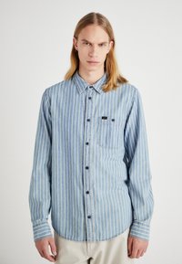 Camicia in denim blu con strisce verticali bianche, design a bottoni, maniche lunghe e una tasca sul petto con una piccola toppa del logo.
