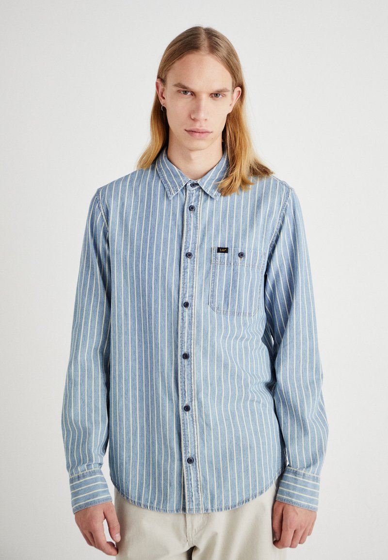 Camicia in denim blu con strisce verticali bianche, design a bottoni, maniche lunghe e una tasca sul petto con una piccola toppa del logo.