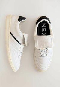 White athletic shoes with a smooth leather finish, black accent on the heel, and a foldable flap over the laces, featuring textured stitching.