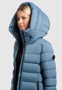 Vaaleansininen toppatakki, jossa on suuri huppu, tikattu pinta, musta vetoketju ja sivutaskujen yksityiskohdat, lisäksi hihassa pyöreä logopatchi.