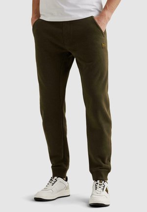 Muž v olivovo zelených teplákoch a bielych teniskách, s rukami v kapsách, stojaci pred jednoduchým pozadím.