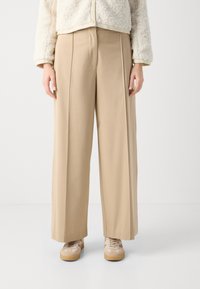 Beige wide-leg trousers with front pleats, made of a smooth fabric. Paired with light-colored sneakers and a fluffy cream jacket.