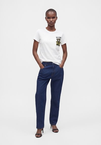 Γυναίκα που φοράει λευκό t-shirt Moschino με στάμπα αρκουδάκι, σκούρο μπλε τζιν και μαύρα πέδιλα με τακούνι, στέκεται με τα χέρια στις τσέπες.