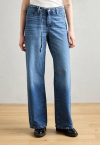 Jean large bleu clair en denim, avec une taille haute, des passants de ceinture, et un léger dégradé. Associé à des chaussures noires.
