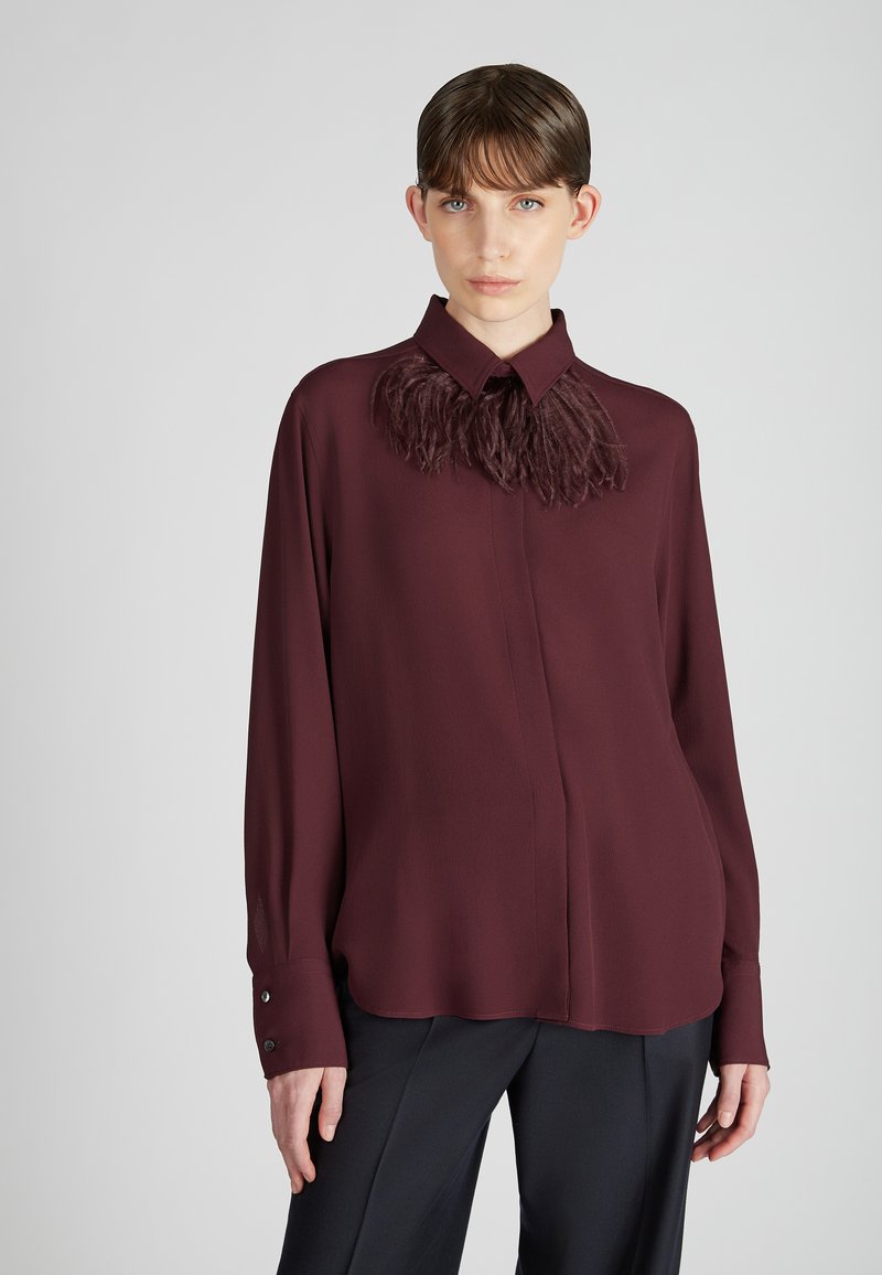 Vrouw met kort bruin haar, gekleed in een lange mouwen bordeauxrode blouse met veerdetails bij de kraag en zwarte broeken, tegen een lichte achtergrond.