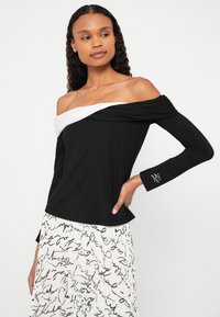 Fekete vállra húzott felső fehér drapériás nyakkivágással, hosszú, ráncolt ujjakkal és az ujjon hímzett díszítéssel.