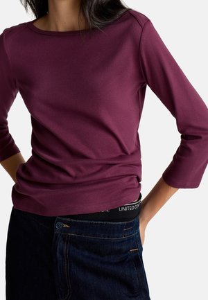 Frau trägt ein langärmeliges, dunkelviolettes Oberteil zusammen mit blauen Jeans und steht mit einer Hand in der Tasche vor einem einfarbigen Hintergrund.