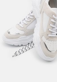 Vita och beige mockasneakers med tjocka sulor och vita snören, en sko på sidan som visar sulan och randiga lösa snören.