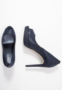 Un paio di scarpe con tacco alto blu navy con una superficie lucida e testurizzata, una mostra la vista superiore e l'altra una vista laterale su uno sfondo bianco.