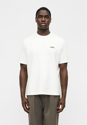 Hombre joven con camiseta blanca lisa de manga corta con un pequeño logo en el pecho, combinada con pantalones marrones de corte holgado, de pie frente a un fondo blanco.