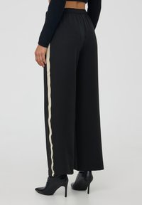 Donna che indossa pantaloni neri a palazzo con strisce beige sui lati e stivaletti neri con tacco alto, in piedi su uno sfondo semplice.