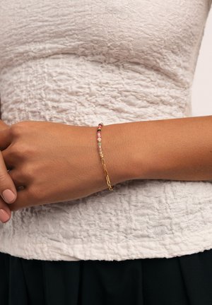 Bracciale in oro con perline piccole assortite in rosa, rosso e nero, indossato su un polso contro uno sfondo bianco texturizzato.