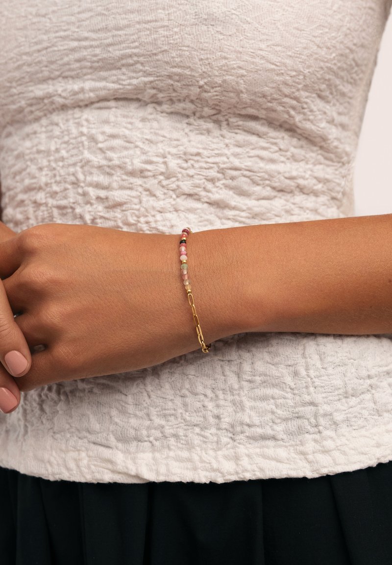Pulsera de cadena de oro con pequeñas cuentas en rosa, rojo y negro, llevada en una muñeca sobre un fondo de top blanco texturizado.