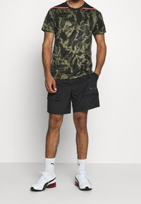 Hombre vistiendo una camiseta de camuflaje verde y negro, pantalones cortos cargo negros con bolsillos, calcetines blancos de Puma y zapatillas blancas con acentos rojos.