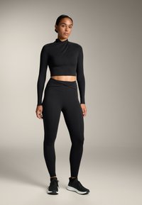 Fekete hosszú ujjú cropped top magas nyakkal; passzoló fekete magas derekú leggings csavart dizájnnal a derékrészen; fekete sportcipők.