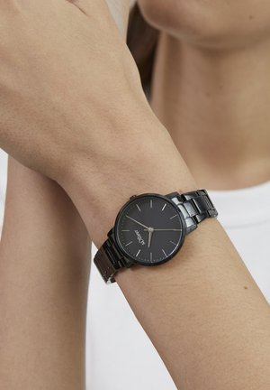 Zwart metalen polshorloge met een ronde wijzerplaat, minimalistisch ontwerp en zilverkleurige accenten; gedragen aan een arm met een lichte huidkleur.