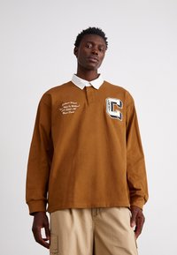 Brun långärmad polo med vit krage, som har en bröstlogotyp och broderade textdetaljer. Avslappnad passform och mjukt bomullsmaterial.