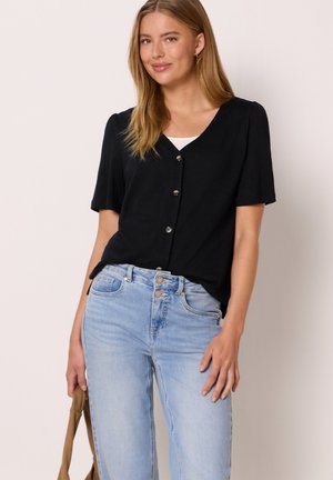 Frau trägt eine schwarze kurzärmelige Bluse mit Knöpfen über einem weißen Top, kombiniert mit hellblauen Jeans mit hoher Taille, und hält eine braune Tasche.