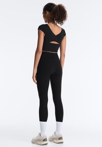 Kvinde med krøllet hår i sort croppet top med udskæring på ryggen og sorte leggings, stående med hvide sokker og beige sneakers.