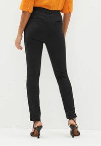 Pantalons noirs ajustés avec une texture lisse, dotés de deux poches arrière, associés à des sandales noires à talons hauts et un blouse orange.