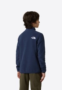 Marineblå fleecejakke med høy hals, med en hvit logo på øvre del av ryggen. Myk tekstur og lange ermer. Kombinert med olivengrønne bukser.