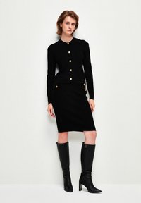 Svart ribbad stickad cardigan med guldfärgade knappar, i kombination med en svart penkjol. Modellen bär knähöga svarta läderstövlar.