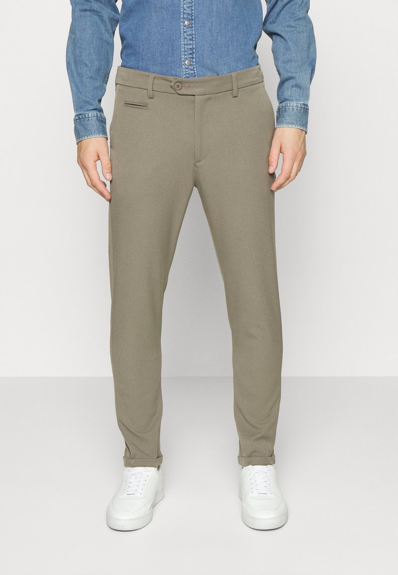 Homme portant un pantalon beige slim fit, retroussé aux chevilles, associé à des baskets blanches et une chemise en denim bleu.