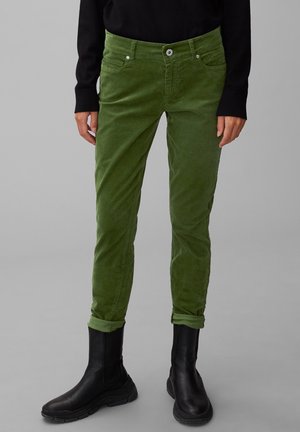 Personne portant un pantalon en velours côtelé vert olive retroussé aux chevilles, un haut noir à manches longues et des bottes noires à semelle épaisse sur un fond gris.
