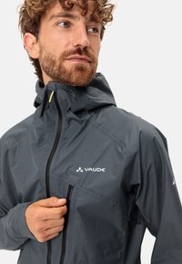 Giacca Vaude grigia con cappuccio, dotata di zip frontale e tasca sul petto. Texture liscia, materiale leggero con logo del marchio sul petto.