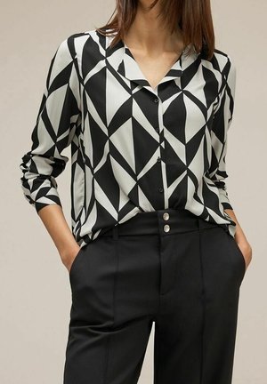 Kobieta ubrana w czarno-białą bluzkę z geometrycznym wzorem na długim rękawie, wsuniętą w czarne spodnie z wysokim stanem i srebrnymi guzikami, z rękami w kieszeniach.