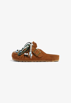 Sandal in suède marrone con dettagli in frange. Presenta una cintura con fibbia e lacci decorativi in perline di diversi colori. Suola in gomma.