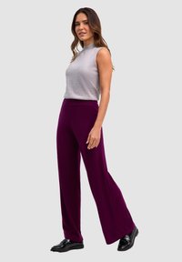 Donna che indossa un top grigio senza maniche e pantaloni larghi viola, in piedi contro uno sfondo semplice grigio chiaro.