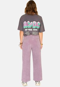 T-shirt graphique AC/DC gris avec manches courtes, vue arrière montrant une impression multicolore de la tournée de 1979 ; associé à un pantalon en velours côtelé large de couleur lavande.