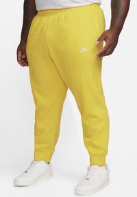 Pantalones de chándal amarillos de Nike con cinturilla elástica, piernas ajustadas y un logo blanco. Combinados con zapatillas blancas de Nike. Textura suave.