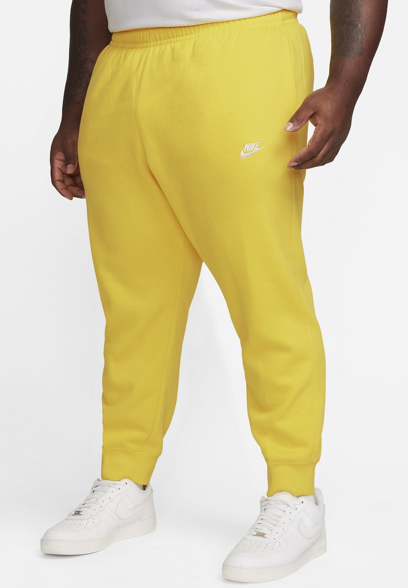 Pantalones de chándal amarillos de Nike con cinturilla elástica, piernas ajustadas y un logo blanco. Combinados con zapatillas blancas de Nike. Textura suave.