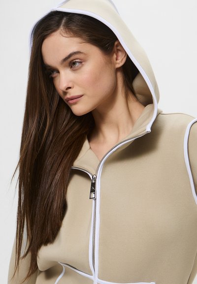 Felpa beige con zip e dettagli bianchi, dotata di cappuccio. Tessuto morbido e dettaglio in metallo argentato sulla zip.
