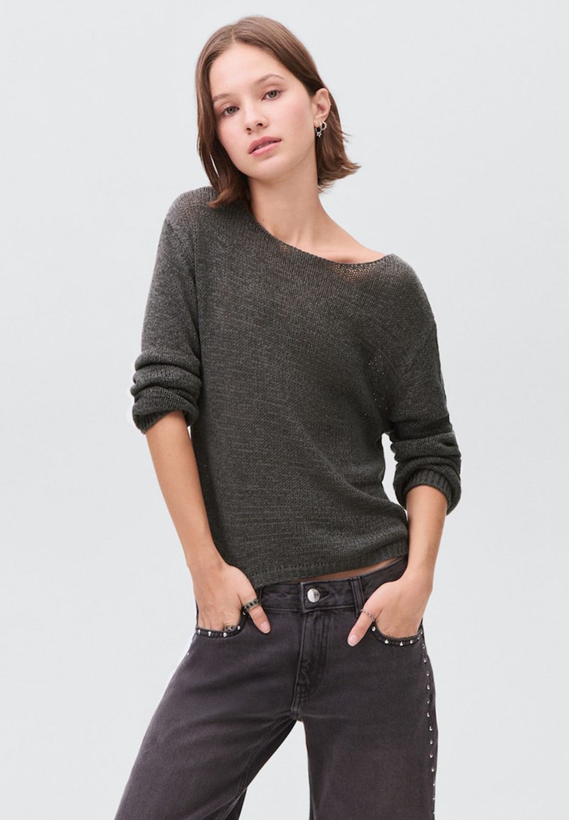 Junge Frau mit kurzen braunen Haaren, die einen dunkelgrauen Off-Shoulder-Pullover und schwarze, mit Nieten besetzte Jeans trägt und die Hände in den Taschen hat.