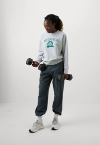 Szara bluza z napisem „MONACO”, ciemnozielone spodnie dresowe, trzymająca czarne hantle, nosząca jasne trampki i białe skarpetki. Gładkie tło.