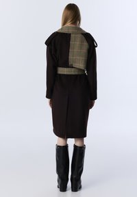 Vrouw staat met haar rug naar ons toe, gekleed in een donkerbruine lange jas met een groene ruitgordel en schouderdetails, gecombineerd met zwarte kniehoge laarzen.