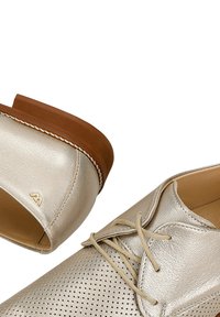 Scarpe in pelle dorata metallica con dettagli perforati, design con lacci e suola marrone simile al legno. Logo accentato vicino al tallone.