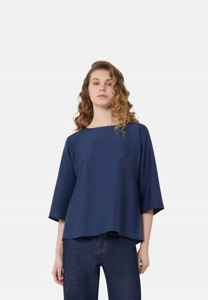 Donna con capelli mossi che indossa una maglia blu navy larga con maniche tre quarti e jeans blu scuro, in piedi davanti a uno sfondo chiaro.